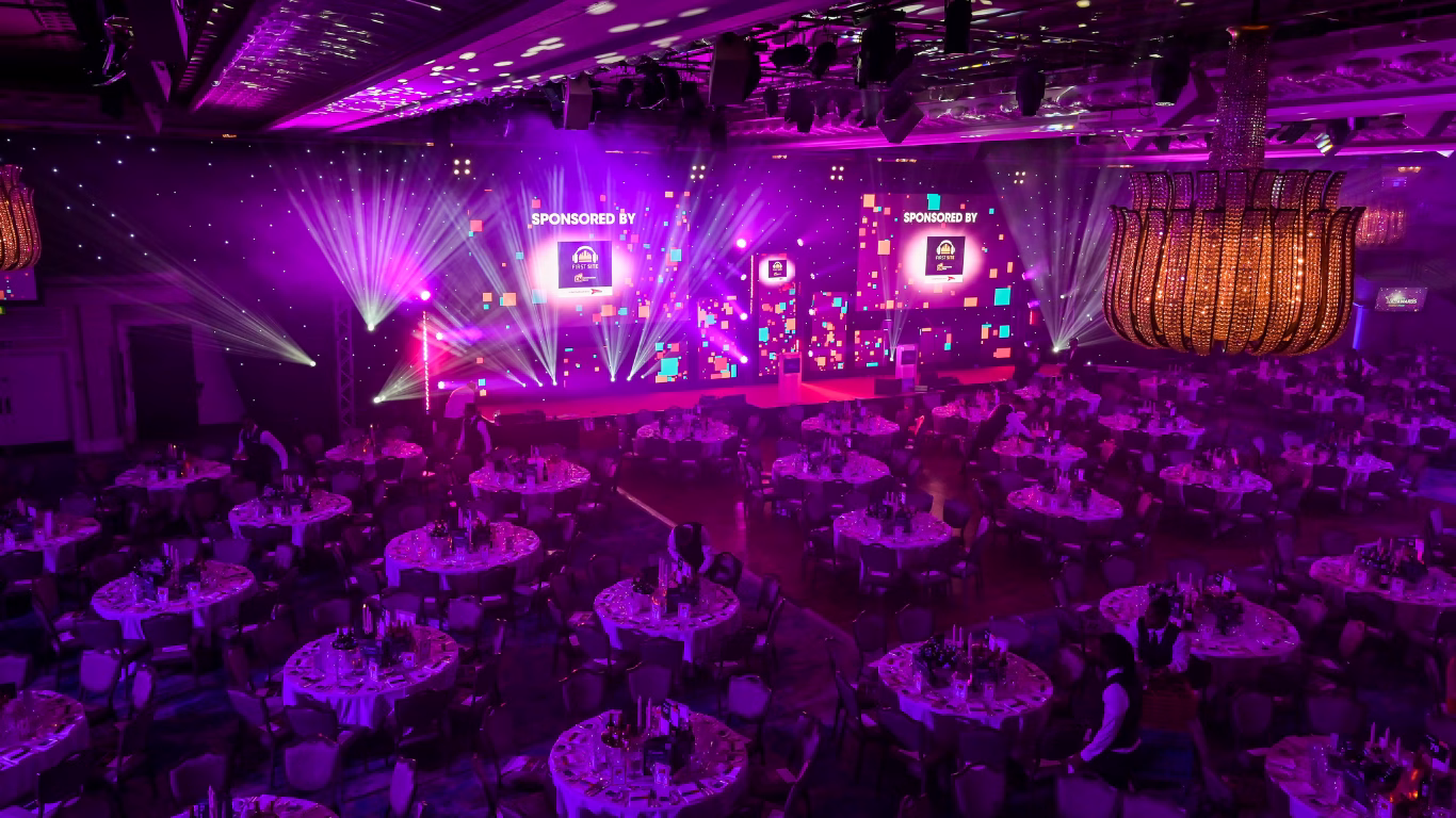Large event venue with multiple tables, a big screen, and purple lighting.